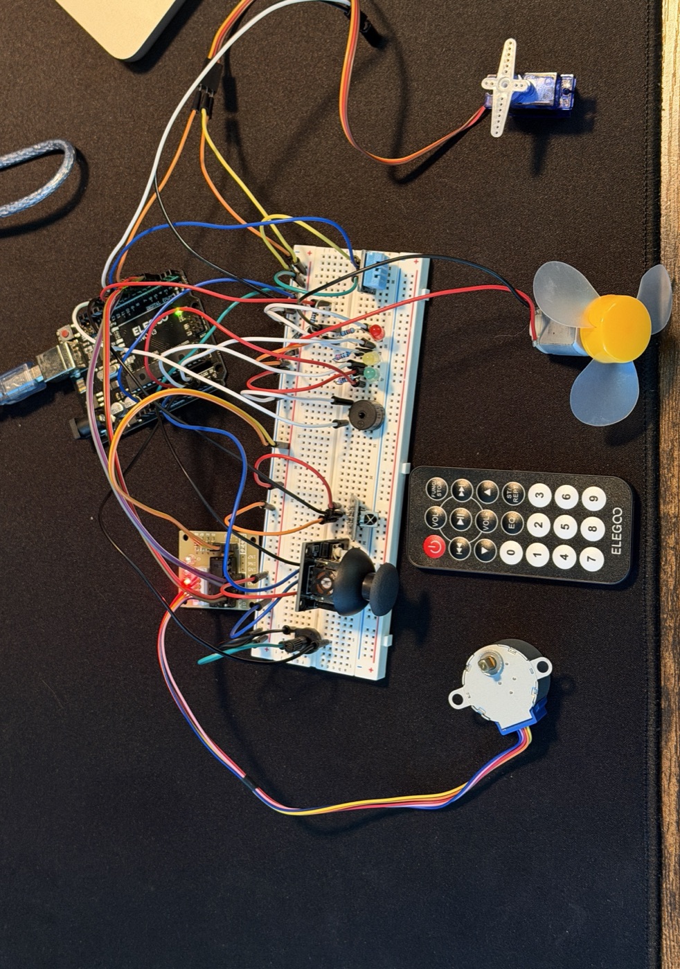 The Arduino test bench: a breadboard carrying status LEDs, a buzzer, a potentiometer, an IR receiver and a joystick, wired to an Arduino board, with a micro servo, a DC fan, an IR remote and a stepper motor with its driver arranged around it.